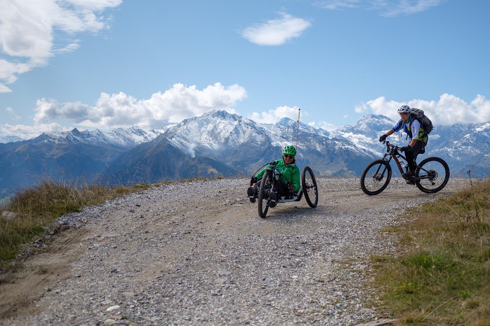Andrea Szabadi-Heine fährt mit einem Handbike auf einem unbefestigten Schotterweg in den Alpen. Hinter ihr ist noch eine Person auf einem Mountainbike zu sehen.
